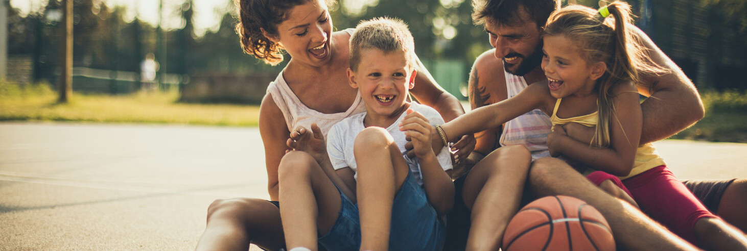 Eine Familie (Frau, Mann, Sohn und Tochter) sitzt auf einem Basketballplatz mit einem Basketball neben ihnen und lachen.