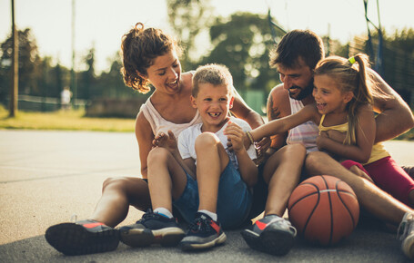 Eine Familie (Frau, Mann, Sohn und Tochter) sitzt auf einem Basketballplatz mit einem Basketball neben ihnen und lachen.