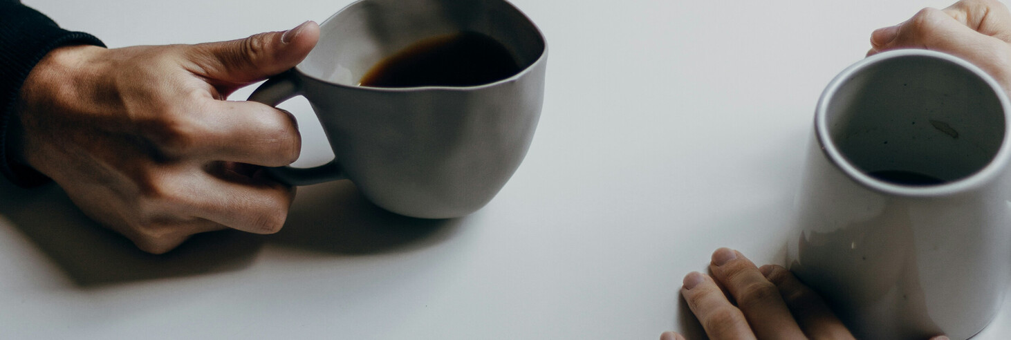 Zwei Personen sitzen an einem weißen Tisch und halten jeweils eine graue Tasse mit Getränk. Man sieht nur die Hände und Tassen.