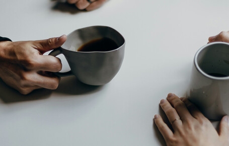 Zwei Personen sitzen an einem weißen Tisch und halten jeweils eine graue Tasse mit Getränk. Man sieht nur die Hände und Tassen.